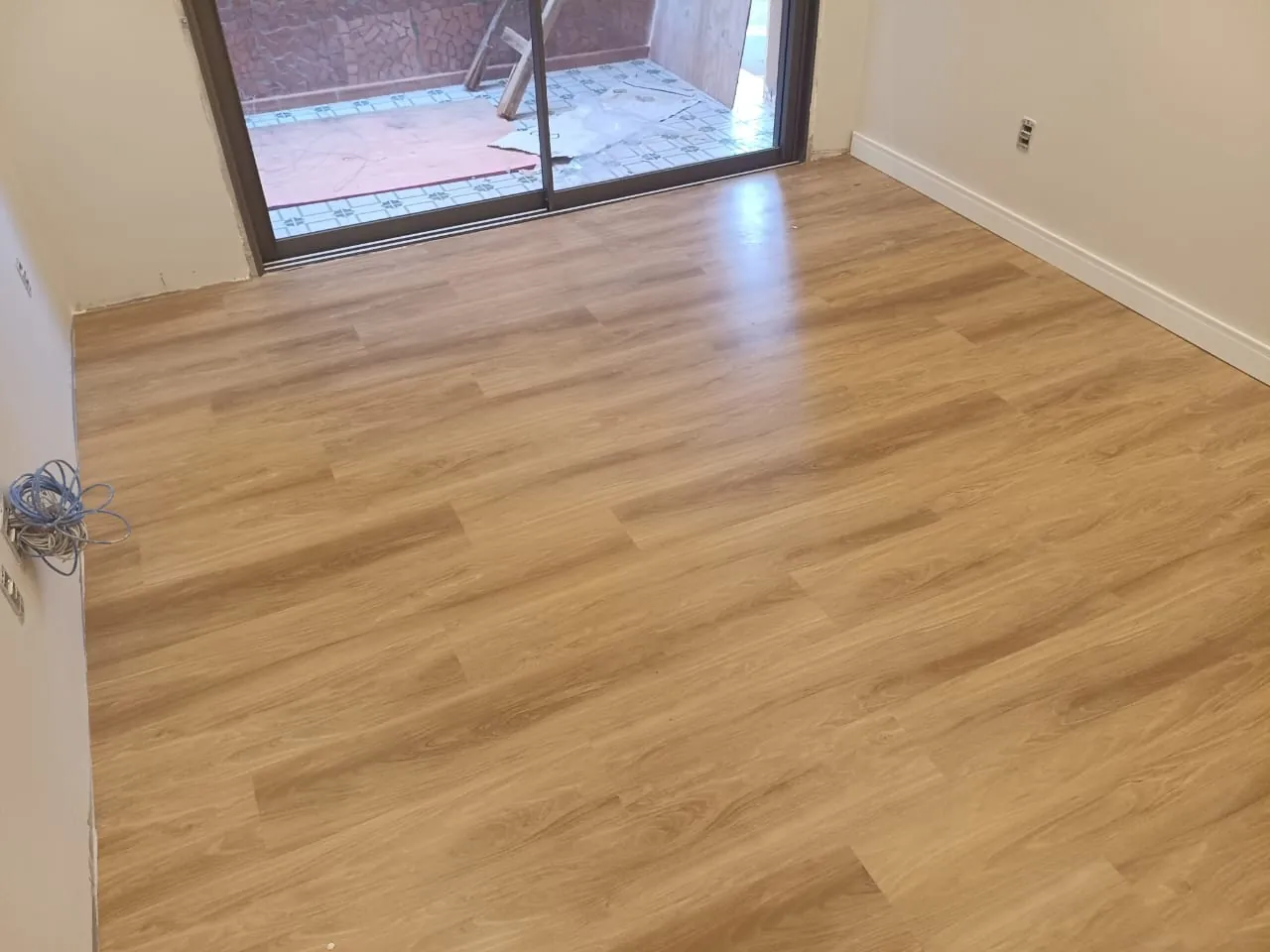 Sala com piso de aspecto amadeirado, rodapés brancos e porta de vidro.